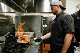 Kitchen employee holding a pan over the stove with flames coming up cooking