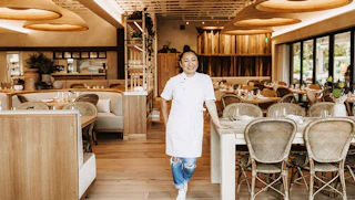A smiling Chef Ann Ahmed in a white apron stands in her Khaluna restaurant with wicker chairs, wooden tables, and soft lighting.
